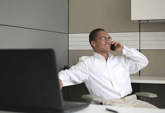 professionally dressed man sitting at desk in front of computer and speaking to client on his phone