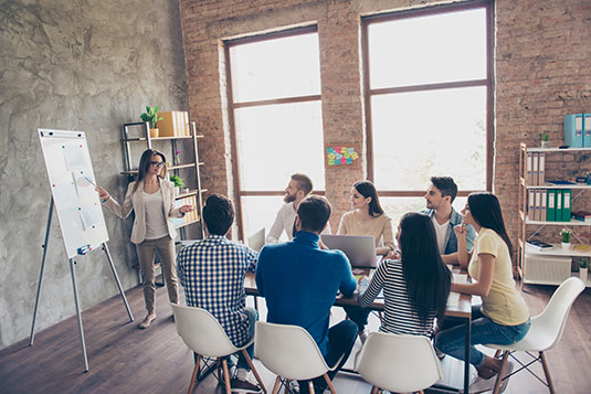 young smart woman in glasses is reporting to the team of colleagues about the new project at the meeting with the white board. Workers are listening to her, all dressed in casual outfits